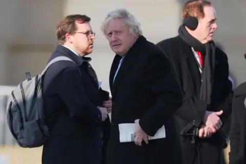 Getty Images Boris Johnson stands outside wearing a coat, flanked by two other men. He is looking at the camera and holding a piece of paper to his left 