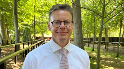 Tom Williams/BBC Andrew Morris standing outside at Newmarket Racecourse. He is standing in a shaded area of the course and there are rows of green leafy trees towering above him. He is looking directly at the camera and smiling and is wearing a white shirt with a pale pink patterned tie.