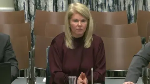 NI Assembly NI Water Chief Executive Sara Venning answering questions from assembly members at Stormont's infrastructure committee.   She has straight, shoulder-length blonde hair and is wearing a loose-fitting maroon crew-neck top or dress.