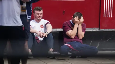 PA Media Two Hearts fans sit on the ground beside a red wall after the match, with one holding his head in his hands in disappointment.