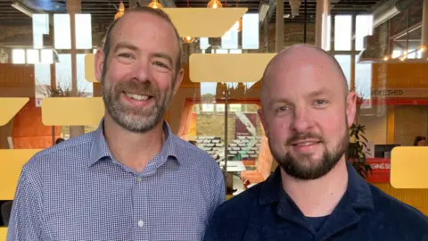 Myles (left) and Giles (right) both standing side by side looking at the camera and smiling. They are wearing shorts and standing in front of a window with colourful designs on it. Myles has short dark hair and facial hair. Giles has a bald head and facial hair.