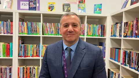 Vikki Irwin/BBC A head and chest image of a man in a library at a school with shelves the book cases with rows of colourful books in the back ground. The head is wearing a blue suit, shirt and tie. He has a purple lanyard on and is smiling at the camera