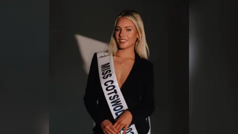 Alice Williams The image shows blonde woman wearing a black long sleeve dress and a white sash across her body with the writing "Miss Cotswold 2025".