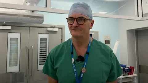 The image shows a man in green scrubs and large glasses is wearing a surgical head covering. He has a blue lanyard around his neck and is holding his hands behind his back. 