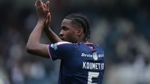 Billy Koumetio in blue Dundee shirt, facing slightly away from camera so you can see his name and number, applauds fans