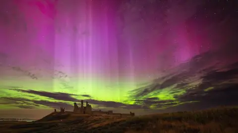 BBC Weather Watchers / Paul Abbleby The night sky filled with bright purple and shafts of white light fills the top of the image while greens of the aurora are seen closer to the horizon where a rolling hill with a ruined castle sits.