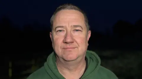 Parkrun volunteer Richard Smith is looking at the camera. He has short hair and is wearing a green hooded top.