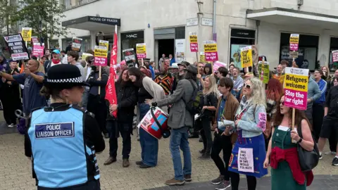 A crowd of people holding signs and looking to the left towards the counter protesters. There is a police liaison officer stood in front of the crowd wearing a blue jacket and a hat.