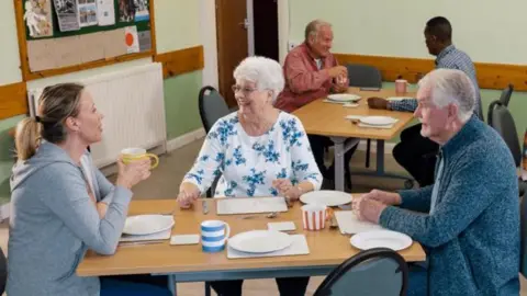 An older man and woman sat at a table next to a younger woman in a community centre. They all have colourful stripey mugs and are smiling.