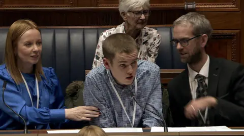 Northern Ireland Assembly Caleb has short brown hair and is wearing a checked-grey shirt and is speaking into a microphone fixed to a bench in front of him. He is sitting between a woman and man and there is another woman sitting behind him.
