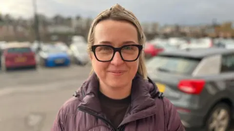 A woman with blonde hair tied back with a scarf is wearing glasses and a purple padded jacket. There are blurred cars parked in the background