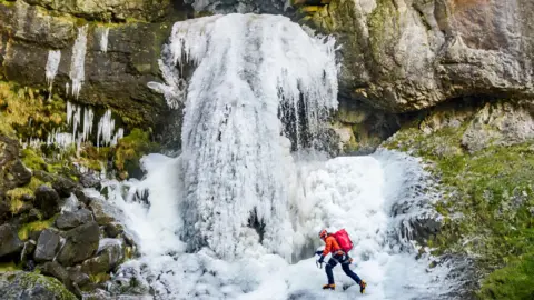 Danny Lawson/PA Wire A climber wearing a helmet, crampons, and a red backpack ascends a frozen waterfall using an ice axe. Thick blue‑white ice sheets cover the rock face, with long icicles hanging from the cliffs above. 
