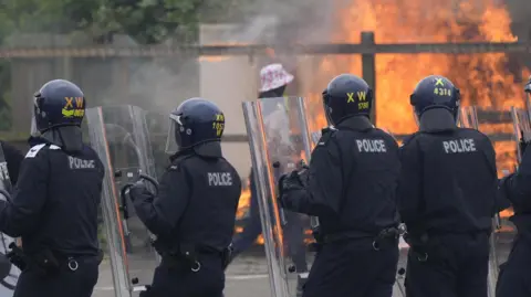 A line of police officers in riot gear in front of a blaze.