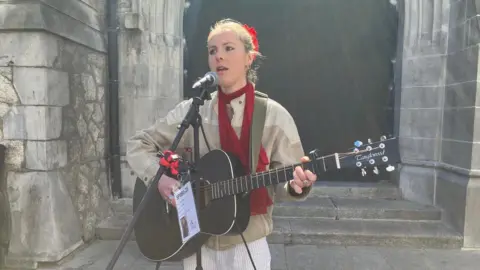 A woman stands singing into a mic and holding a guitar. has blonde hair, scraped back from her face in a red bobble. She wears a white top and a skinny red scarf.