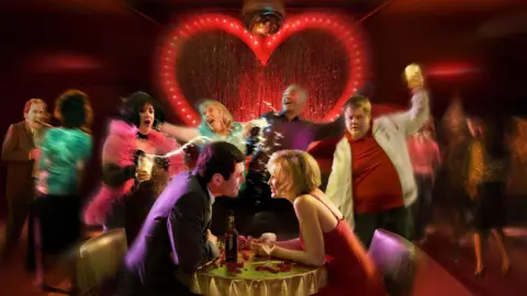 Gavin and Stacey at a table sharing a drink with the rest of the characters dancing behind them