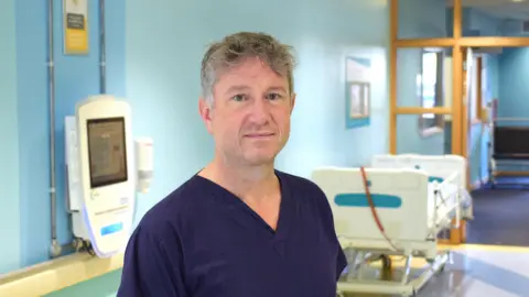 University of Southampton Professor of Paediatric Surgery Nigel Hall stood in a hospital ward wearing dark blue scrubs