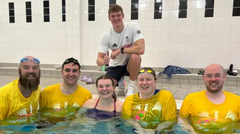 Oliver Morgan is crouching on the side of a swimming pool wearing a white T-shirt. Below him in the pool are five people. Two men on the left are wearing yellow Children in Need T-shirts, a woman is wearing a swimming costume. To her right is a woman and a man, both also wearing yellow Children in Need T-shirts