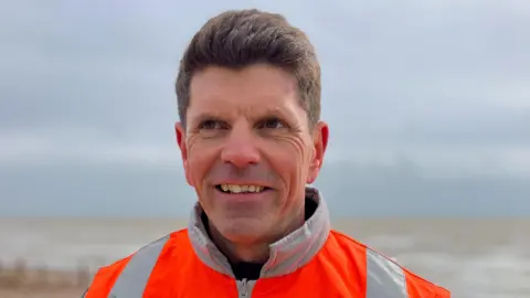Phil Cresswell-Nash, a man in an orange hi-viz jacket, looks away from a camera.