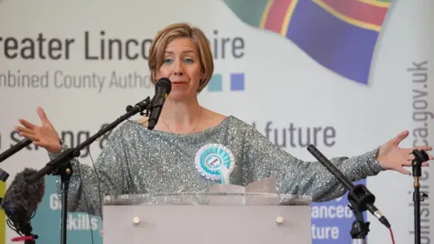Dame Andrea Jenkyns stands at a podium surrounded by four black microphones. She has her arms open and is wearing a sparkly silver dress with a blue and white rosette.