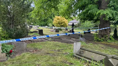 White and blue police tape surrounds a number of stone grave among trees in a churchyard. 