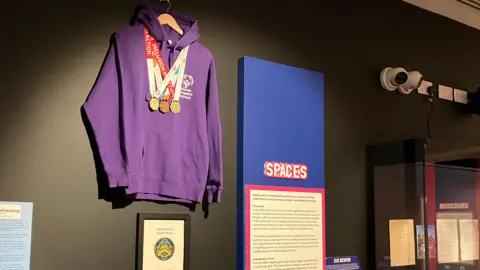 Jon Wright/BBC A museum exhibition with a purple hoodie on a coat hanger with three medals round the neck. There are text labels on the wall as well.