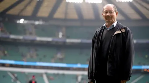 PA Media Ian Robertson is standing inside a rugby stadium which is blurred in the background. He has a bald head and glasses and is wearing a black jacket over a dark jumper and a light-coloured shirt.