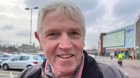 George is looking at the camera and is wearing a brown leather jacket with a pink collar poking out from underneath.