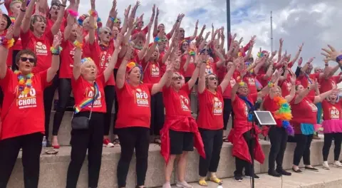 Members of the All For One choir wearing red T-shirts with their arms raised while singing in the open air