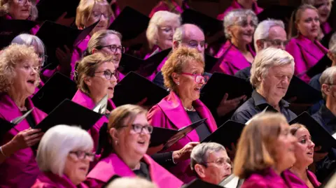 A choir full of women and a few men. The women all have pink jackets on. Everybody is looking straight ahead while singing.