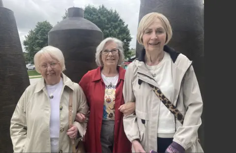 Shows three women in fair hair standing in front of a public artwork
