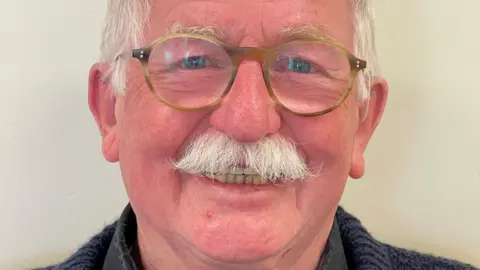 Gloucestershire County Council Councillor Bernie Fisher. He is smiling. He has white hair and a moustache. He wears rounded glasses, and a navy knitted jumper.