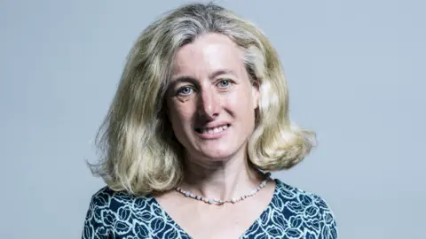 Woman with short blonde hair and a patterned blue top and beaded necklace stands infront of grey background 