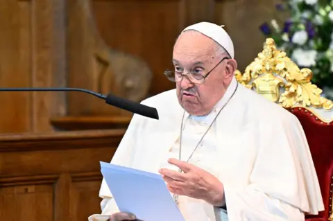 Pope Francis sitting in a velvet chair, reading from a sheet of paper. He is speaking into a microphone. The background is blurred.