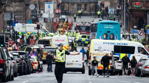 Getty Images A busy city street filled with emergency vehicles, including multiple police cars, ambulances and fire engines, with numerous officers and responders managing a large incident scene. Pedestrians are gathered at the edges while traffic is stopped in both directions.