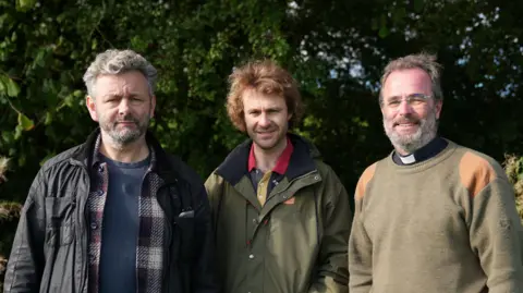 Three men sending side by side. First man has short grey hair and is wearing a blue t shirt, checked shirt and black jacket. The second man is wearing a khaki coloured jacket and the third man - a reverend - is wearing a khaki coloured jumper