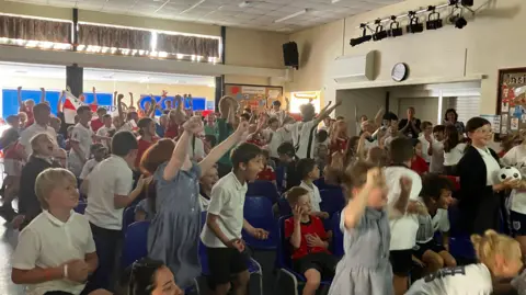 A room full of school children cheer in delight while watching the England Mexico match in a school hall.