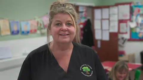 Heather Street - A woman with a black shirt on with the logo of a tree and the name of the nursery in a white font. She looks at the camera and has her blonde hair tied up. It is a head and shoulders shot.