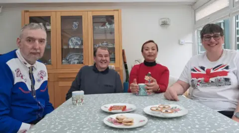 Four people sit around a dining table laden with biscuits and cups of tea.