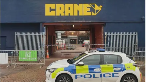 West Midlands Police A police car parked outside a blue building with a square archway. The word crane is in yellow capital letters in the centre of the building