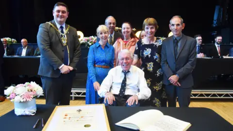 Norman Moorsom, who is seated behind a desk bearing a framed certificate, smiles into the camera. Behind, from left to right, are Middlesbrough Mayor Chris Cooke, Mr Moorsom's friend Linda Ford, his son Richard Moorsom, granddaughter Laura Moorsom, daughter Hilary Webb and son-in-law Martin Webb.