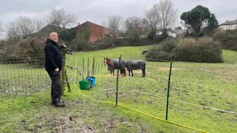 Rob Cheesley stands in front of a field with an electric fence. Three horses can be seen grazing in the background. They are wearing rugs. Cheesley is wearing a black hoodie and trousers. Houses are visible in the distance.