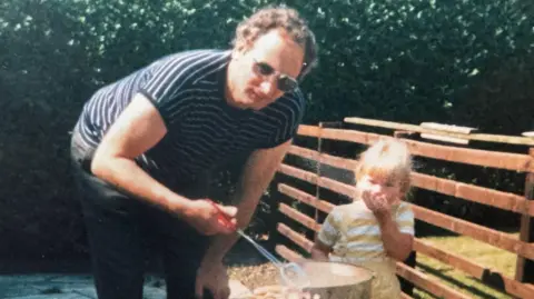 Handout An old photo of Hubert Dodds cooking on a barbeque. He is leaning over with a hand on his knees and the other is holding silver tongs. He has brown, curly hair and is wearing a blue and white striped t-shirt and dark trousers. There is a little girl beside him with blonde hair who is wearing a yellow and white striped outfit. She is holding her hand up to her face like she is eating something. There are standing to the left of a brown fence and there is a large hedge behind them.