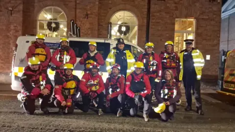 West Mercia Police Eleven people are wearing yellow helmets and red life jackets, standing in front of a police van. It is nighttime. With them are two police officers, wearing yellow hi-vis jackets and black police hats