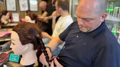 Dominic Murphy is sitting on the right holding a long side plait of a mannequin. head with long brown hair. He has aa bald head and is looking down a the hair, with a hair tie held in his mouth. There are other men also styling with mannequins in the background to the left.