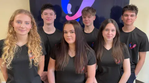 The six-strong team of sixth-formers are standing in front of a screen in two rows, three girls in front and three boys at the back. They are all wearing matching black t-shirts sporting the pink-and-blue logo of Team Valoris.