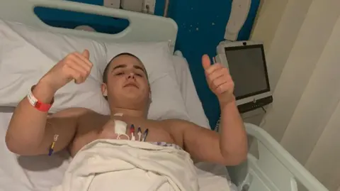 Charlie Taylor, a man with short brown hair, is at the centre of the image. He is lying on a hospital bed and is putting his thumbs up at the camera. There are wires on his chest. 