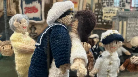 Knitted figures replicating a scene from World War Two. It shows a sailor in navy uniform dancing with a woman in a white dress. Behind it is a grey-haired woman in a yellow dress who is smiling, and a child in white and blue who is also smiling.
