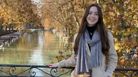 Darcie James Darcie James standing on a bridge overlooking a canal in France, on her year abroad. She has long brown hair and is wearing a coat and scarf and smiling at the camera.