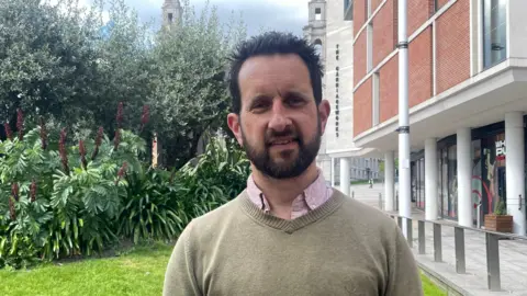 Sofía Luis-Hobbs/BBC A man with dark hair and a dark beard looks at the camera. He wears a sage green jumper on top of a light pink collared shirt. To the left behind him is a mix of greenery, and to the right is a building and paved area.