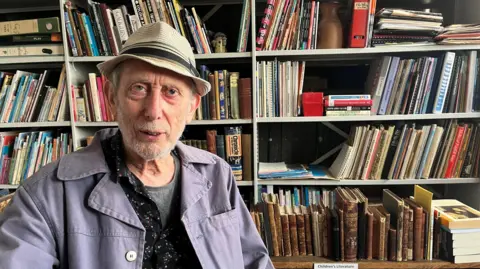 Michael Rosen is pictured successful  his bureau   near  of frame, with shelves of books down  him. He wears a achromatic  chapeau  with a acheronian  set  astir   it and a lilac jacket.